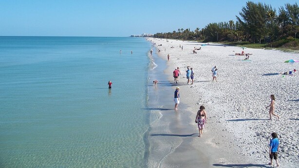 Ein Strand mit klarem Wasser, Menschen die entlanggehen und Palmen.