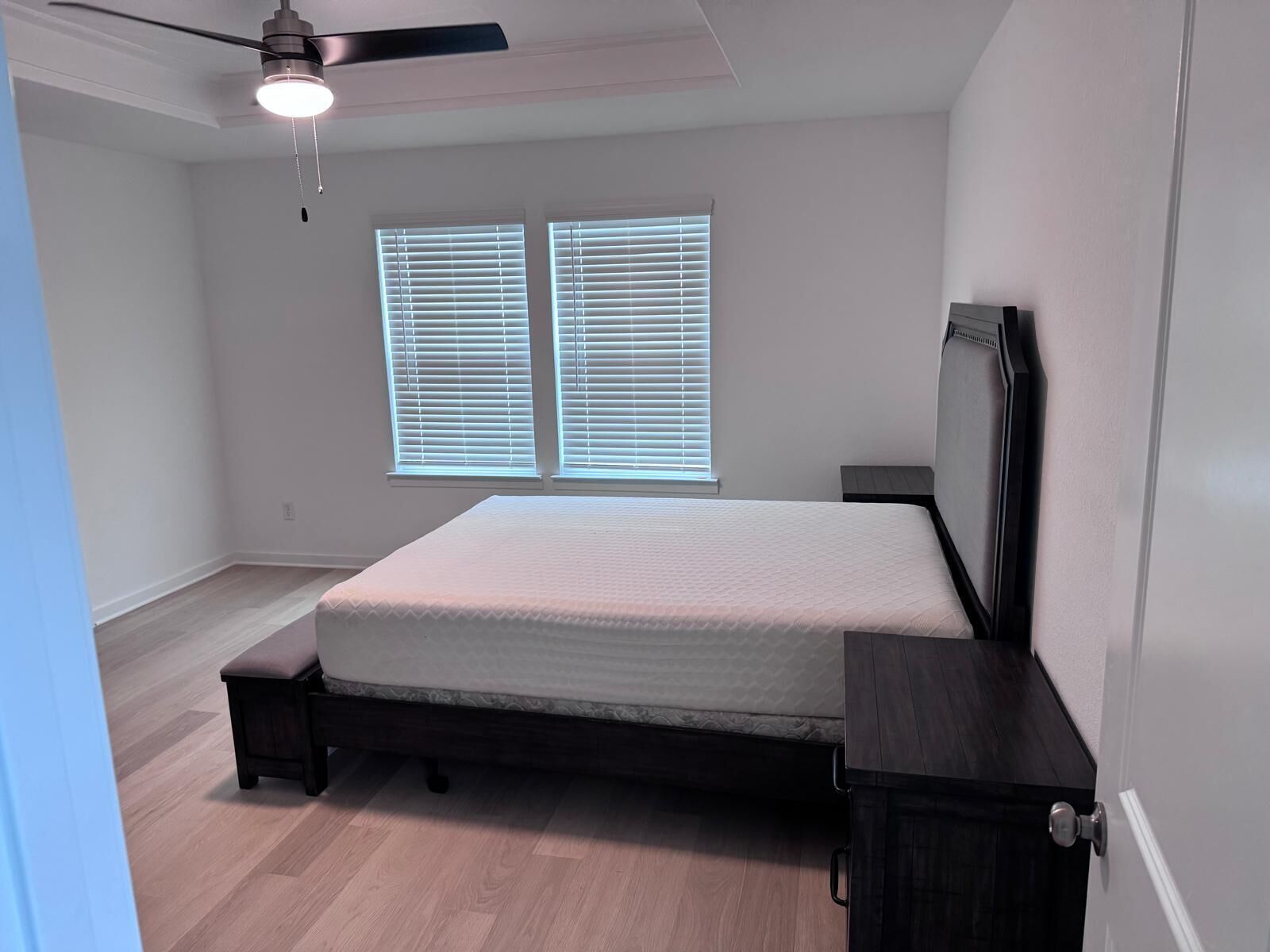 Une chambre à coucher comprenant un lit, un ventilateur de plafond, deux fenêtres avec stores, une table de chevet et une porte.