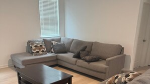 Ein Wohnzimmer mit einem Ecksofa, einem Couchtisch und einem Fenster mit Jalousien.