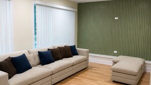 Een woonkamer met een beige bank, een poef, houten vloeren en groen verticaal paneel op de muur.