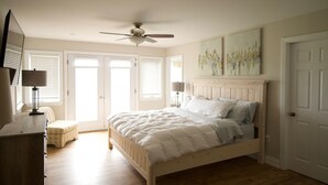 Un dormitorio con cama, ventilador de techo, mesita de noche, lámpara, ventana con persianas y puerta.