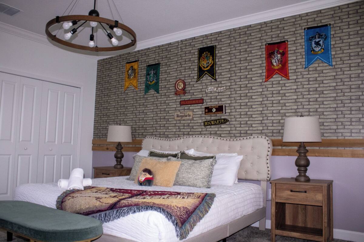 A bedroom with a brick-patterned wall, a bed with multiple pillows, a bench, and a nightstand with a lamp.