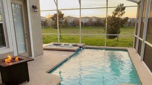 A covered outdoor swimming pool with a fire feature, surrounded by a house with a view of a grassy area and other houses.