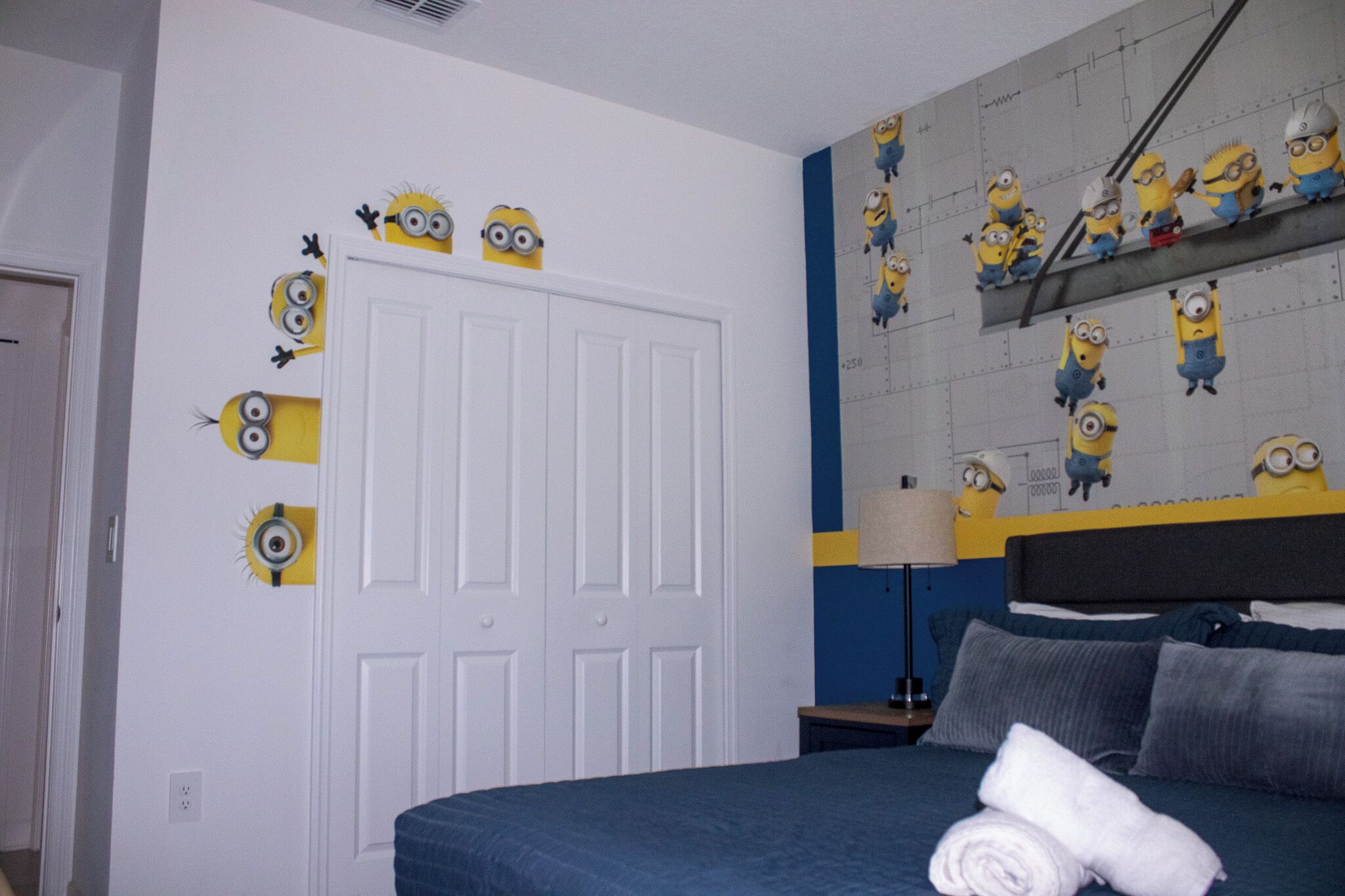 A bedroom with a bed, pillows, a rolled towel, a nightstand, a lamp, and a wall with minion decals.