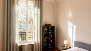 A bedroom with a bed, a nightstand, a lamp, a radiator, a window with curtains, and a shelf with items. - Room in private villa, charm, comfort, near Velodrome & beaches-Private parking lot (Marseille)