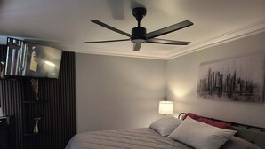 Ett hotellrum med en säng, en takfläkt, en väggmonterad TV, en lampa och en tavla som föreställer en stadsbild.