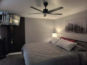 A hotel room with a bed, a ceiling fan, a wall-mounted TV, a lamp, and a cityscape painting. - 1-bedroom condo in delightful, Arcadia area, Phoenix, AZ w/AC (Phoenix)
