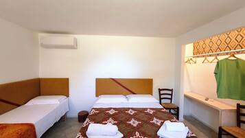 Una habitación con dos camas individuales, una cama grande con colcha estampada, una silla, un taburete pequeño, un perchero con ropa y un aire acondicionado instalado en la pared.