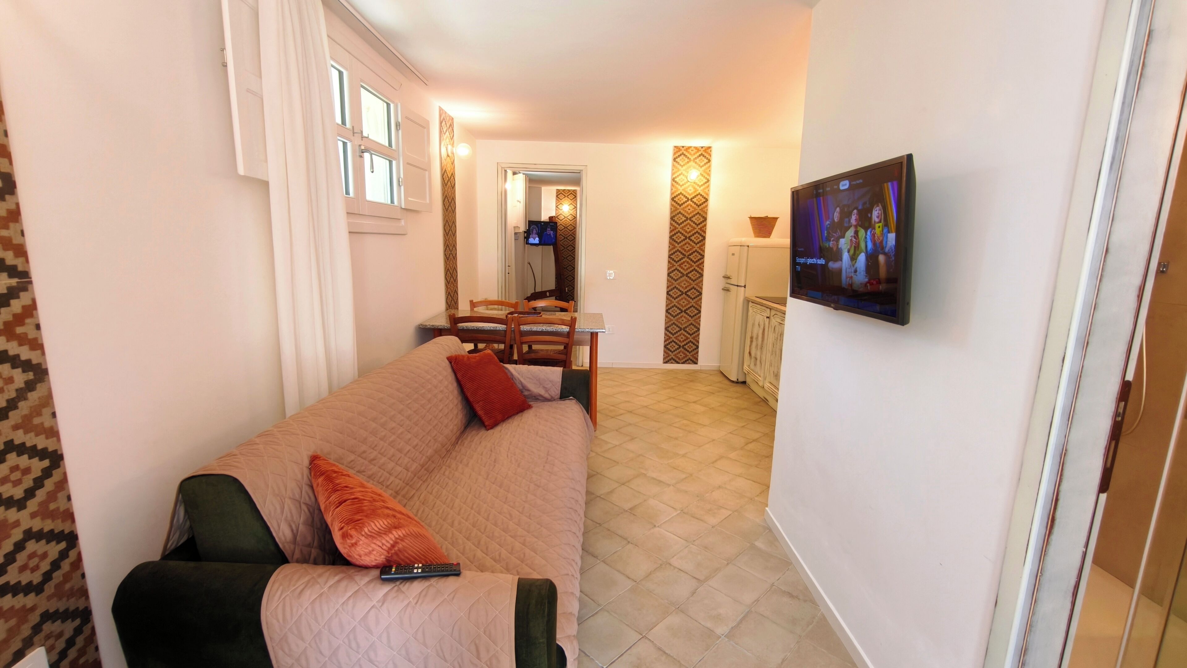 A living room with a sofa, a remote control, a dining area with chairs and a table, and a TV mounted on the wall.