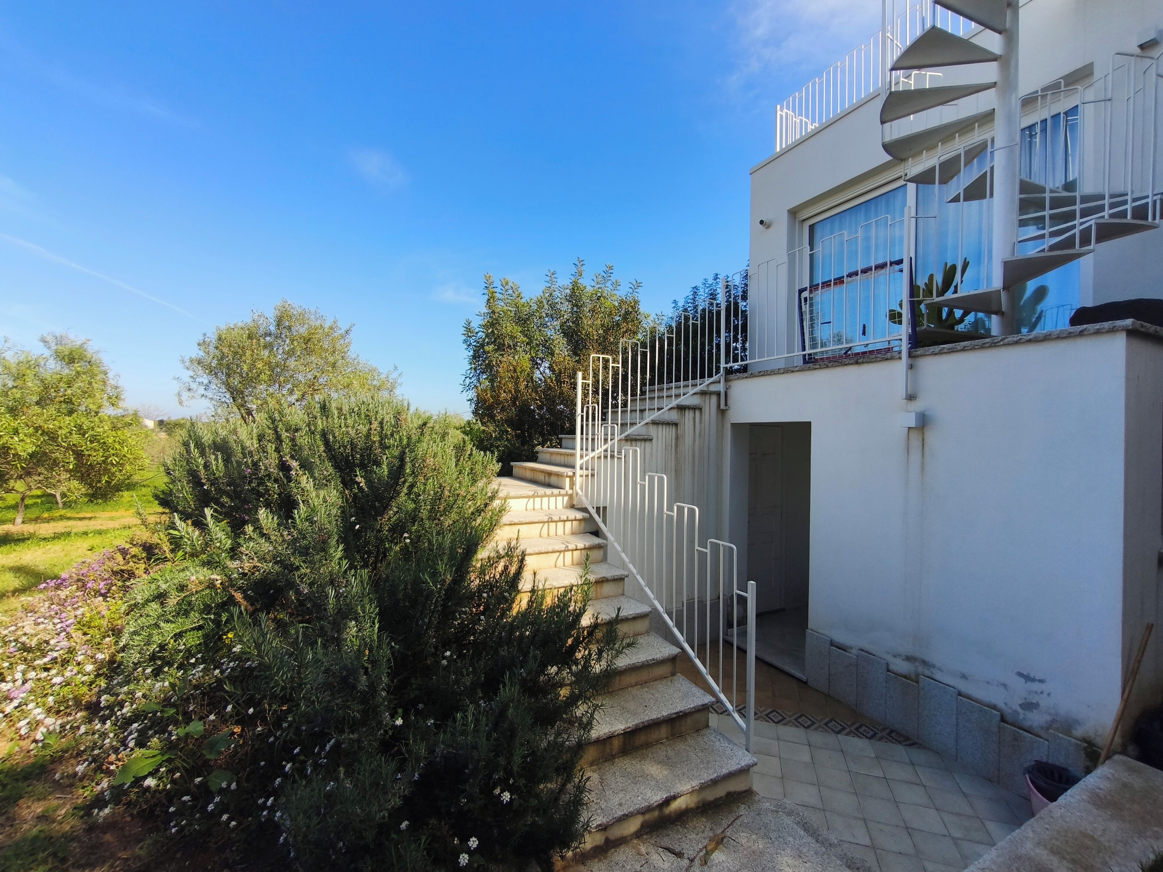 Ein modernes Gebäude mit einer Wendeltreppe, die zu einem Balkon führt, umgeben von Grünpflanzen und blauem Himmel.