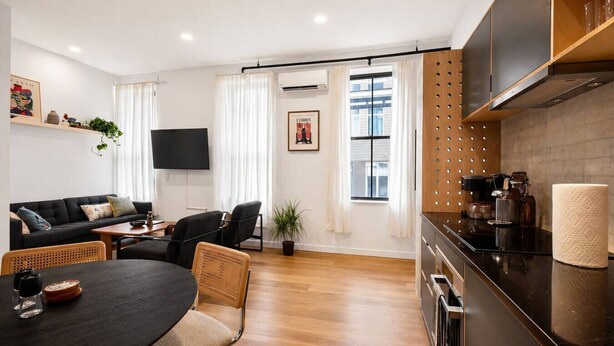 En moderne stue med sofa, spisebord, stole, et fladskærms-tv og et køkkenområde med komfur og skabe.