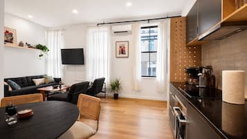 A modern living room with a sofa, dining table, chairs, a flat-screen TV, and a kitchen area with a stove and cabinets.
