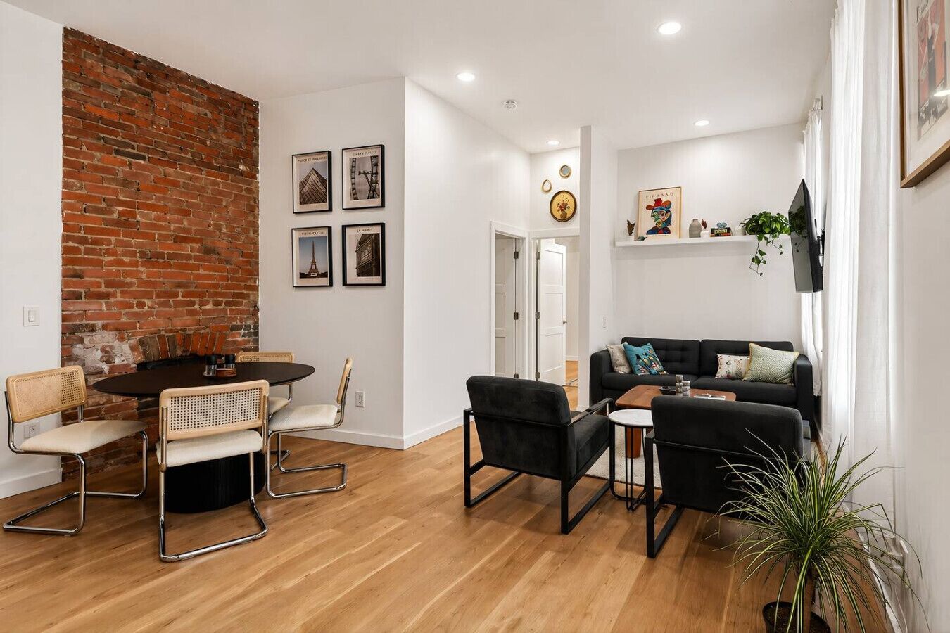 A living room with a brick wall, a dining area with a round table and chairs, a black sofa, and a TV mounted on the wall.