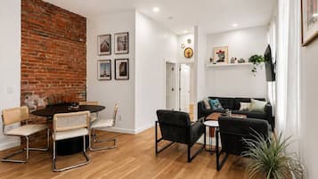 A living room with a brick wall, a dining area with a round table and chairs, a black sofa, and a TV mounted on the wall.