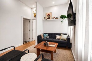 Een woonkamer met een zwarte bank, een houten salontafel en een zwarte fauteuil. Er is een witte deur, een plant en een schilderij aan de muur.