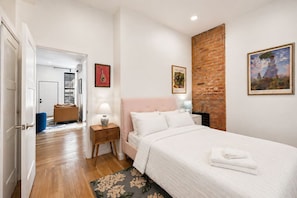 A bedroom with a bed, a nightstand, a lamp, a blue ottoman, a door, a hallway, and a brick wall. - Washington Park 2BR w Parking - Walk to All (Cincinnati)