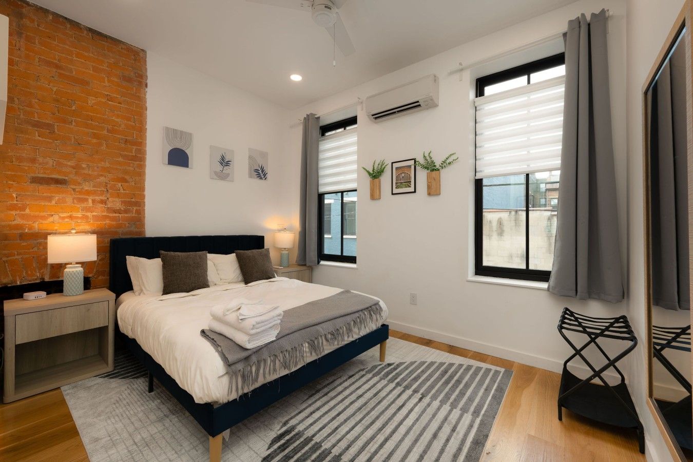 A modern bedroom with a brick accent wall, a bed with a dark headboard, a bedside table with a lamp, and a view of the outside through large windows.