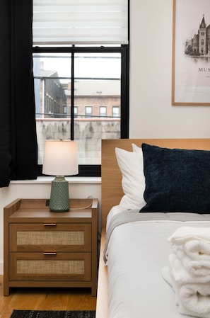 A hotel room with a bed, a nightstand with a lamp, a window with blinds, and a framed picture on the wall. - Washington Park 2BR w Parking - Walk to All (Cincinnati)