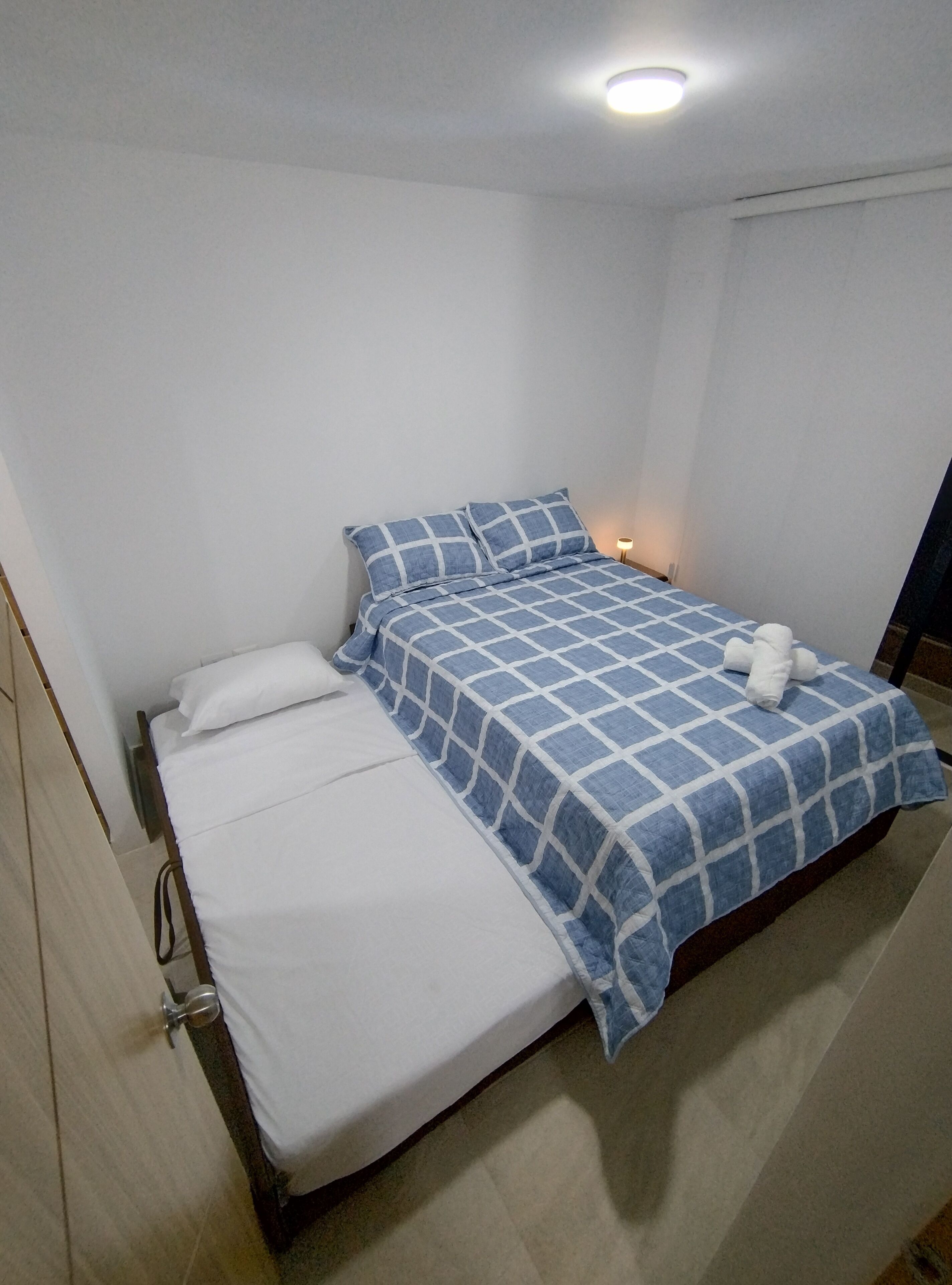 A single bed with a blue and white checkered bedspread, white pillows, and a bedside lamp.
