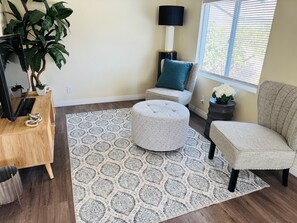 Ein Wohnzimmer mit einem gemusterten Teppich, einem Sofa, einem Hocker, einem Beistelltisch mit Lampe und einer Zimmerpflanze.