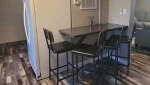 Une cuisine compacte comprenant un réfrigérateur, une petite table à manger, quatre chaises, une plante en pot et une fenêtre avec des stores.