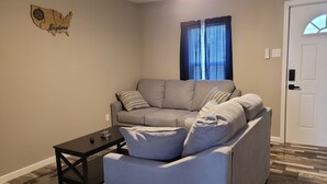 Un salon avec un canapé gris, une petite table basse, un tapis, une décoration murale en bois et une porte.
