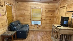 L’intérieur d’un chalet en bois est aménagé avec un canapé en cuir, une petite table, un téléviseur posé sur un meuble en bois et une fenêtre équipée de stores.