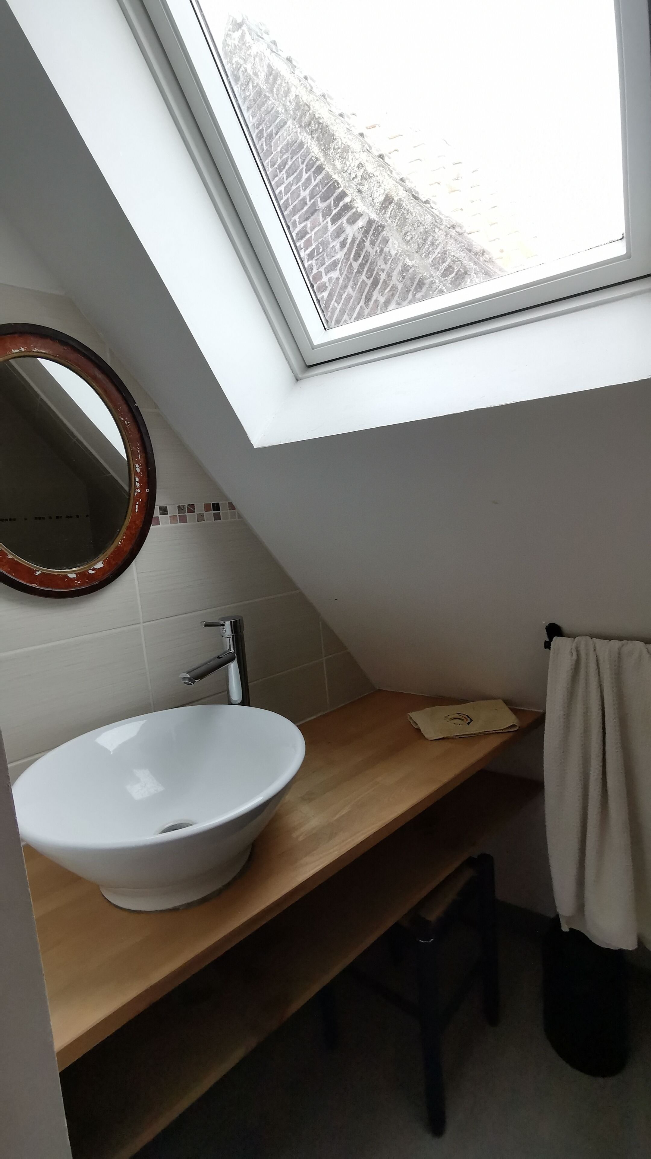 A skylight above a white sink with a wooden countertop, a round mirror, and a towel rack.