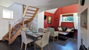 A dining area with a wooden staircase, a dining table set for four, and a living area with a sofa and a window. - La Cense du pont. Gîte in square farmhouse. Lille and Tournai campaign. (Bachy)