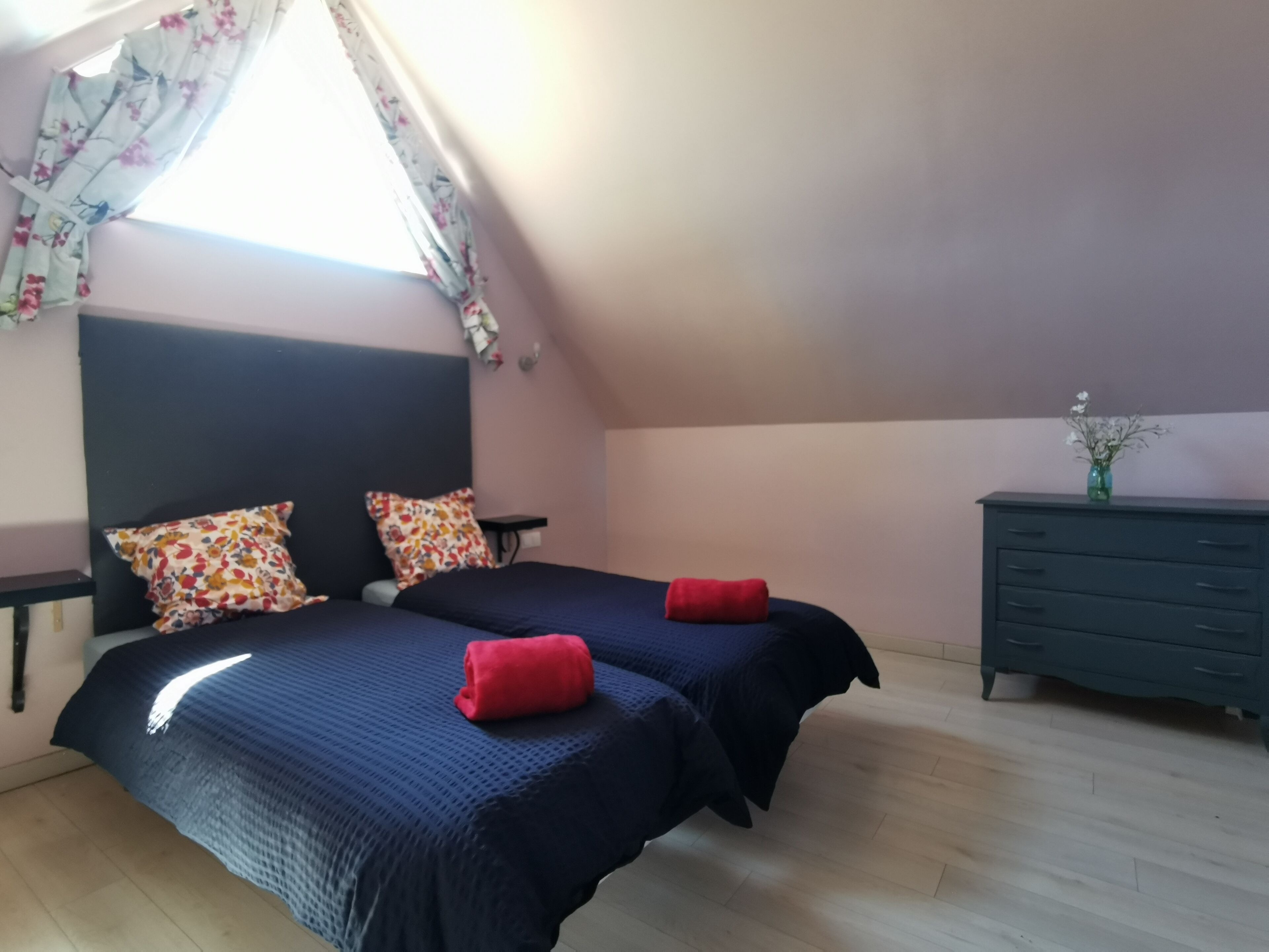 Een slaapkamer met twee bedden, een kaptafel, een raam met bloemen gordijnen en een vaas met bloemen op de kaptafel.