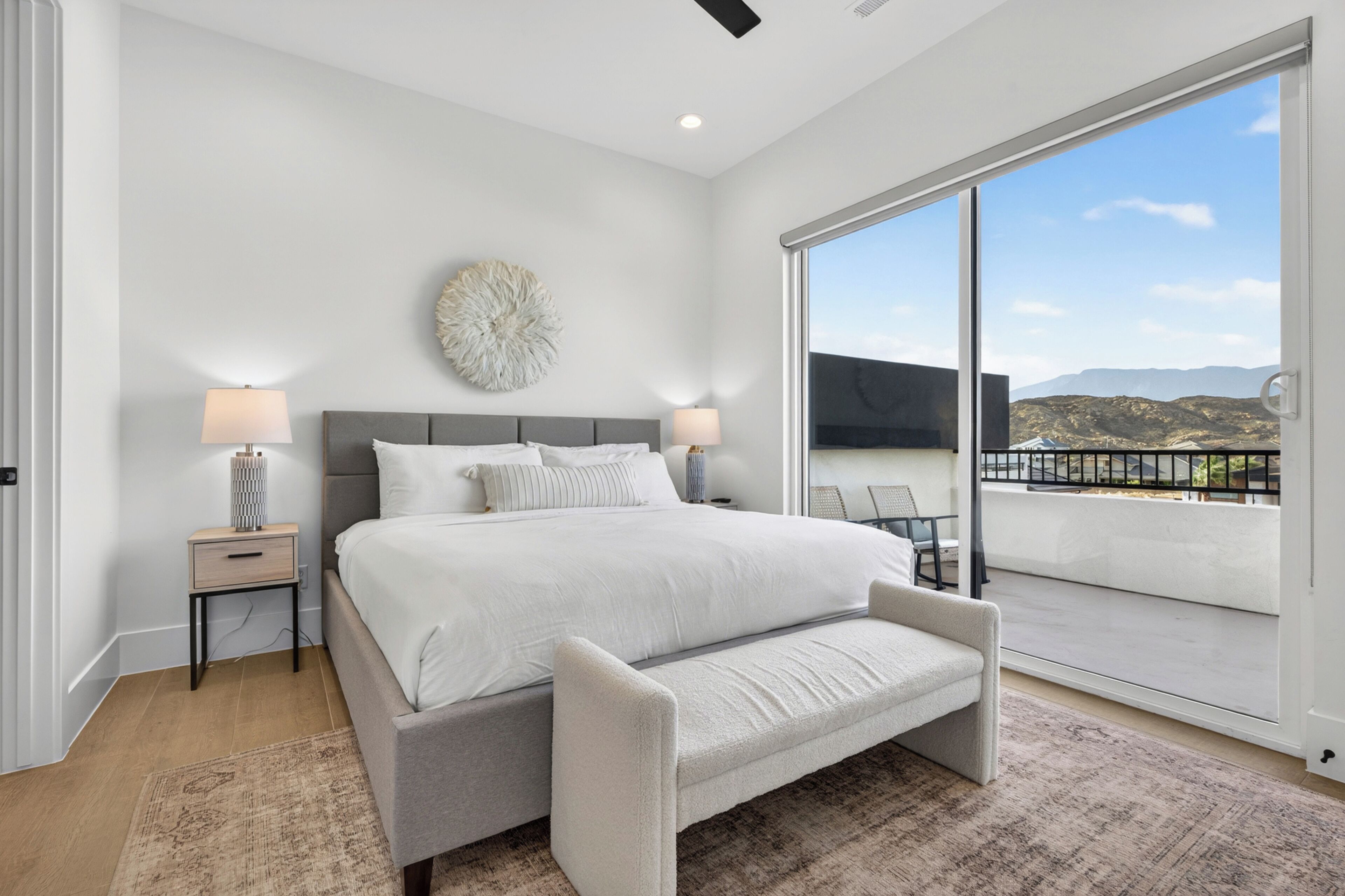 A bedroom with a large bed, bedside tables, a bench, a decorative wall piece, and a balcony with a view.