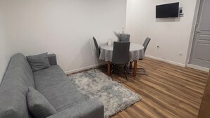 Un salon avec un canapé gris, une petite table ronde entourée de chaises, un téléviseur fixé au mur et un plancher en bois.