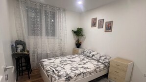 Une chambre à coucher comprenant un lit, un bureau, une chaise, une commode, une plante et une fenêtre avec des rideaux.