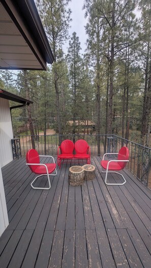 Une terrasse dotée d’un ensemble de meubles de jardin avec un canapé rouge et deux tabourets en bois, entourée d’une rambarde en métal et offrant une vue sur une forêt.