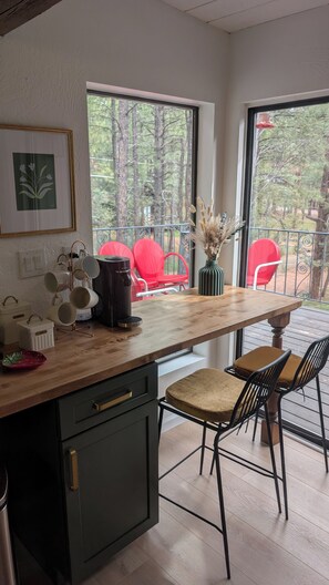 Une cuisine avec un plan de travail en bois, une machine à café et un vase contenant des plantes séchées. Dehors, il y a deux chaises rouges et une table.