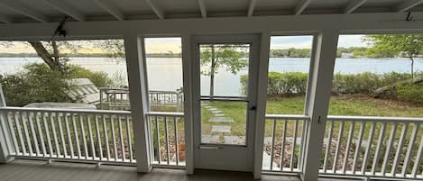 A covered porch with a view of a lake and a house across the water.