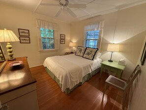 A bedroom with a bed, two bedside tables, a desk, a chair, a ceiling fan, and two windows with shutters. - Charming 4-bedroom cottage on Lake Wequaquet (Barnstable)