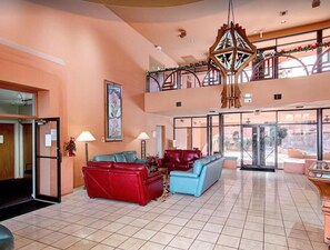 A spacious lobby with a red and blue sofa set, a large chandelier, and a view of the outdoor area through large windows. - CASA GINA - Anasazi Village - Pool View (Phoenix)