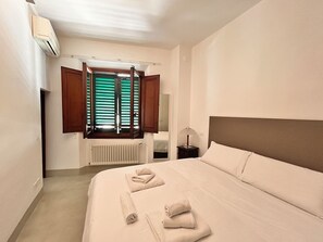 A hotel room with a bed, a nightstand, a lamp, a window with wooden shutters, and an air conditioning unit. - Casa Aligi by FlorenceHospitality (Firenze)