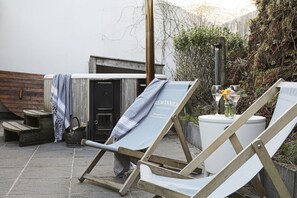 Eine Holzterrasse mit zwei Liegestühlen, einem kleinen Tisch mit Blumenvase und einer Tür, an der ein Handtuch hängt.