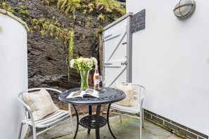 En liten sittplats utomhus med ett bord, två stolar, en vas med blommor och en flaska vin.