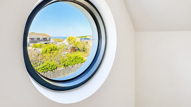 A circular window with a clear view of a residential area, including houses and greenery. - Well Cottage - Three Bedroom Cottage, Sleeps 6 (Trevarrian)