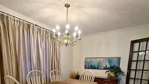 Ein Esszimmer mit Holztisch, weißen Stühlen, einer Chandelierleuchtre, einem Gemälde und einem fenster mit Vorhängen.