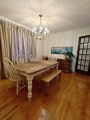 A dining room with a wooden table, white chairs, a chandelier, a painting, and a window with curtains. - Cozy 3 Bedroom Home in Omaha (Omaha)