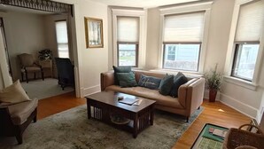 Ein Wohnzimmer mit einer Couch, einem Couchtisch und einem Sessel. Es gibt Fenster mit Jalousien, einen Deckenventilator und eine Tür zu einem anderen Raum.