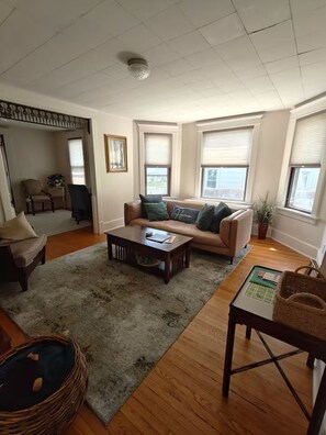 A living room with a sofa, coffee table, and a chair. There are windows with blinds, a ceiling fan, and a door leading to another room. - Cozy 3 Bedroom Home in Omaha (Omaha)