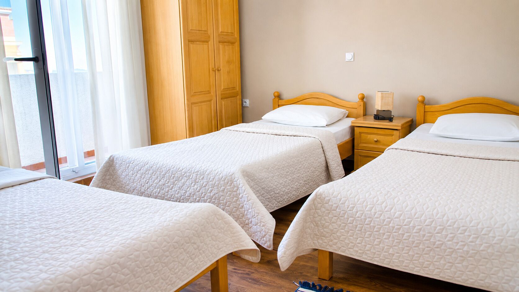 A room with two beds, a wooden wardrobe, a bedside table with a lamp, and a window with curtains.