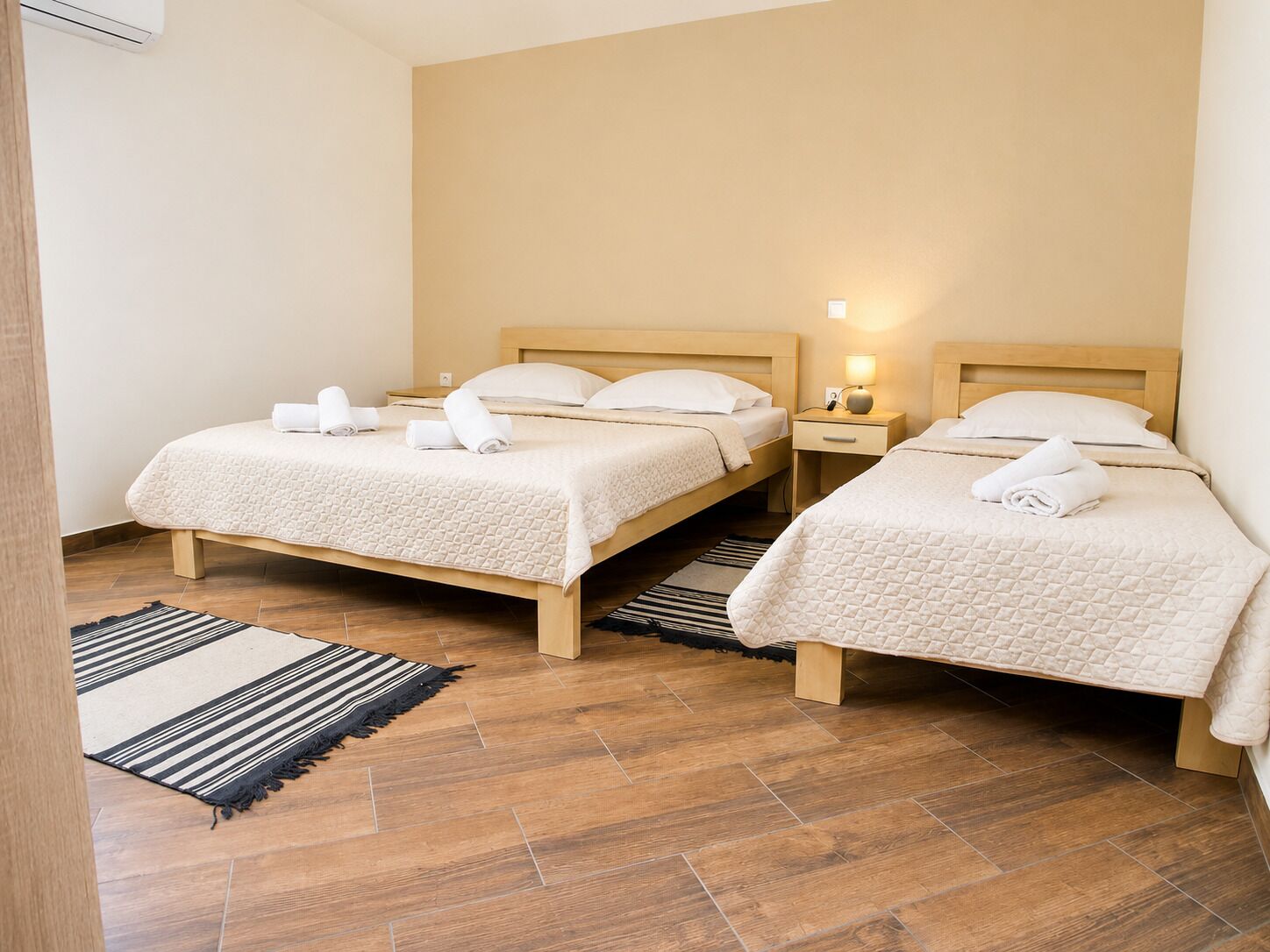 A hotel room with two beds, a wooden floor, a striped rug, and a nightstand with a lamp.