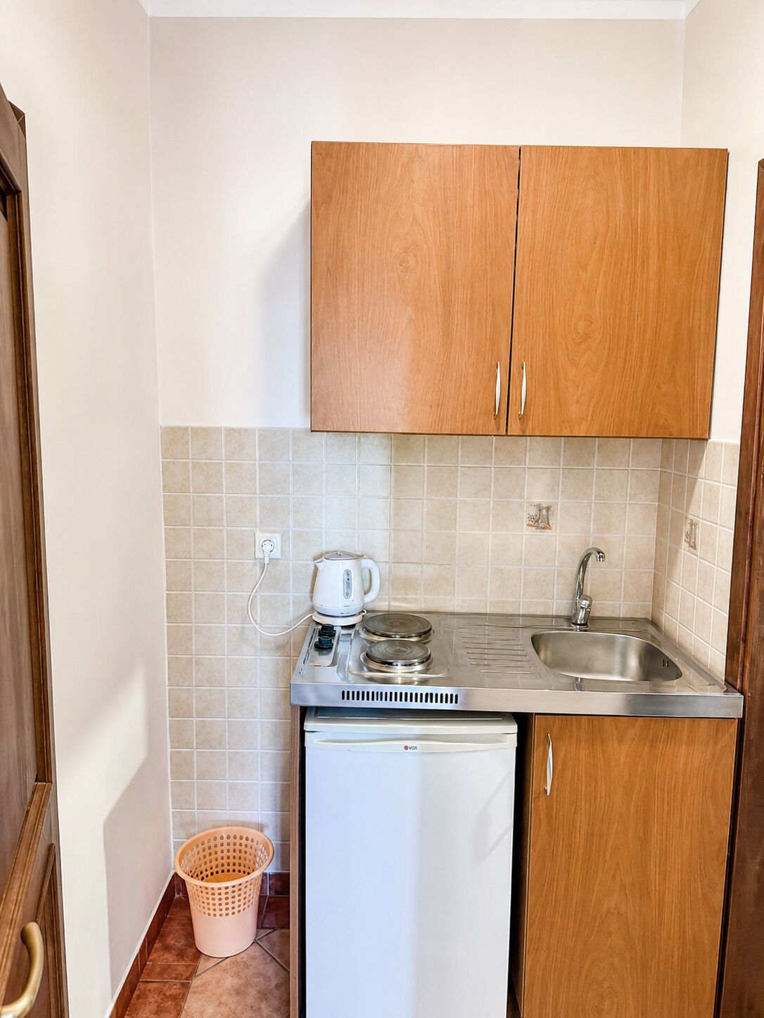 Cocina compacta con armarios de madera, un lavamanos de acero inoxidable, un refrigerador blanco pequeño y una tetera sobre la encimera.