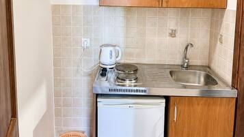 Cocina compacta con armarios de madera, un lavamanos de acero inoxidable, un refrigerador blanco pequeño y una tetera sobre la encimera.